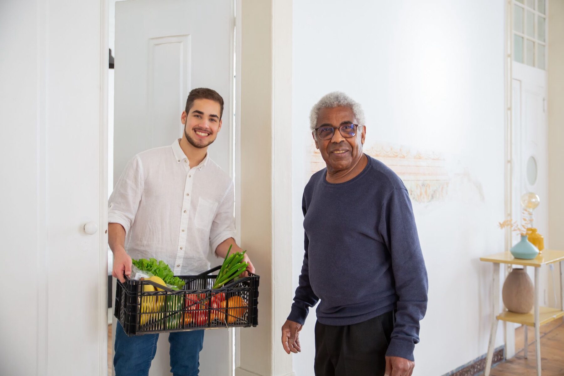 Young man handing a crate of fresh vegetables to an older man at a bright doorway