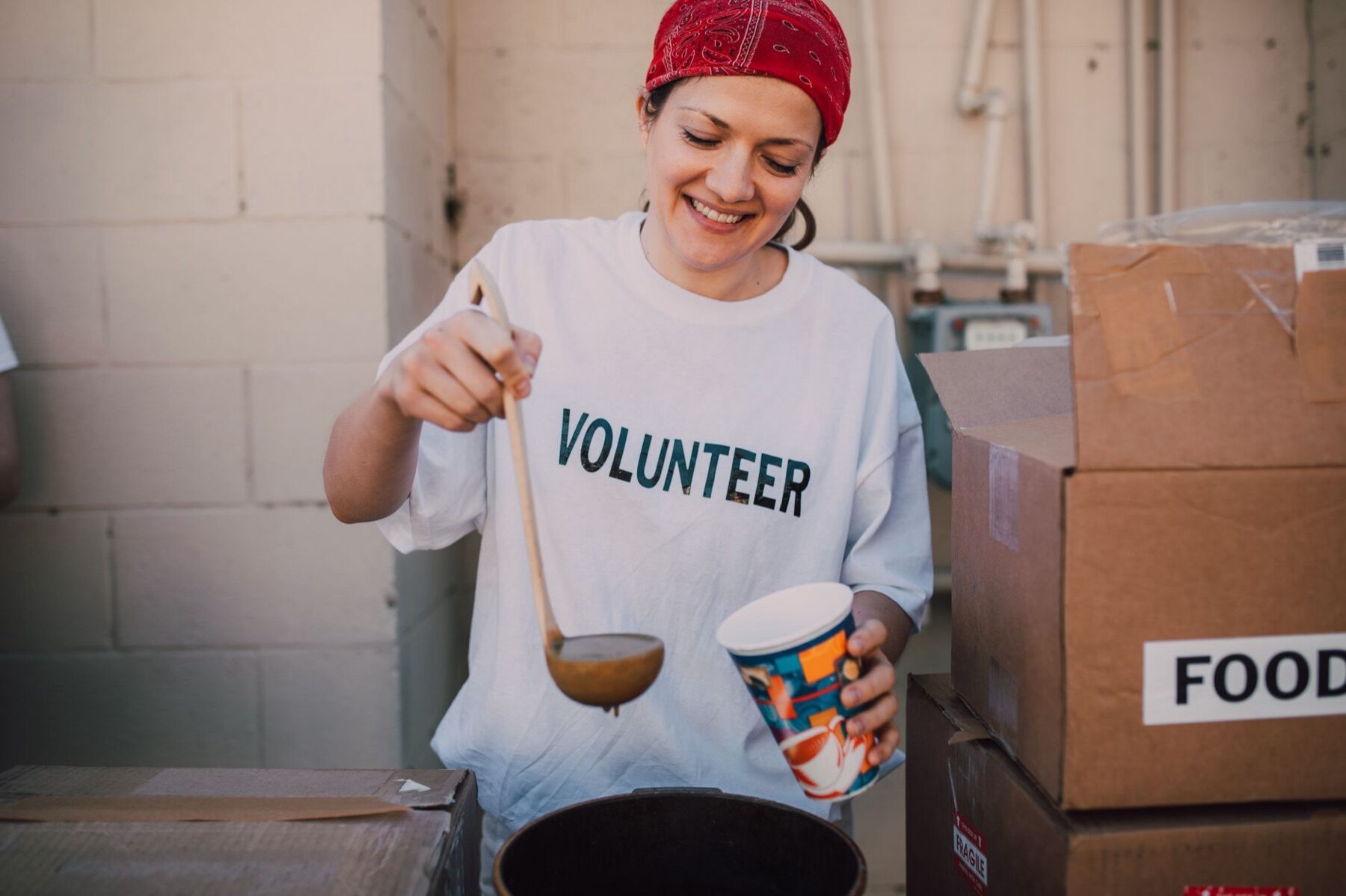 Smiling volunteer in a red bandana ladling soup into a cup at an outdoor food distribution