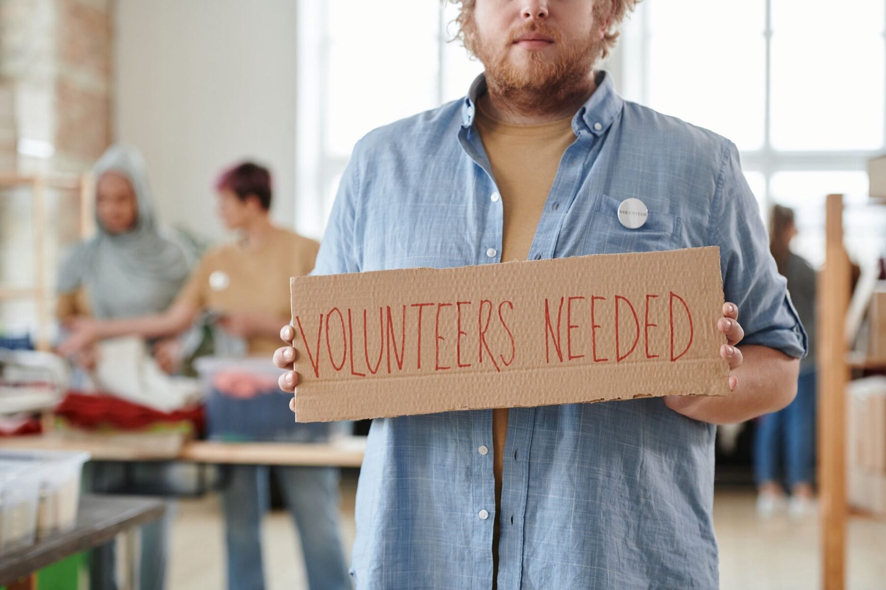 Volunteer holding a hand-lettered cardboard sign reading Volunteers Needed at a donation sorting center