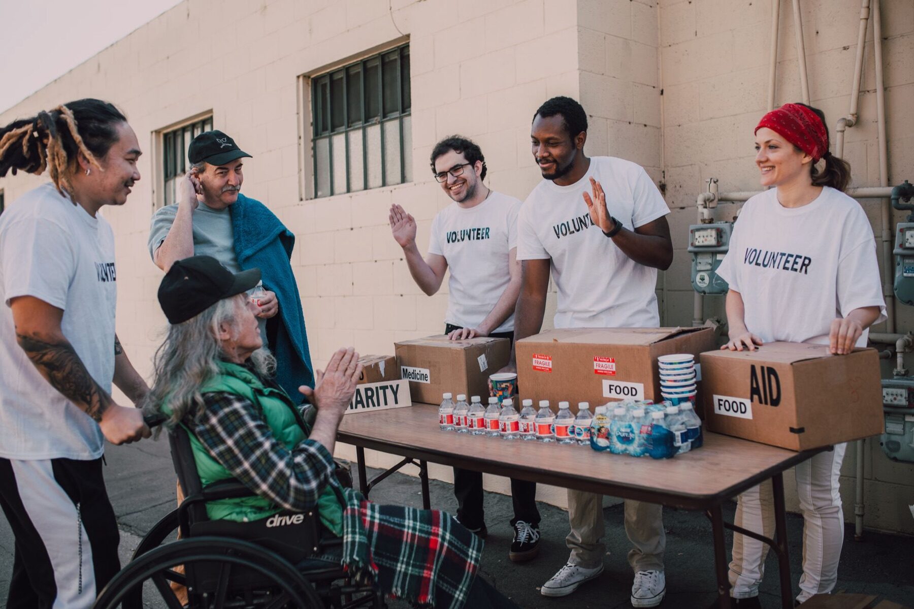 Group of volunteers laughing and waving with an elderly man in a wheelchair at an outdoor distribution table